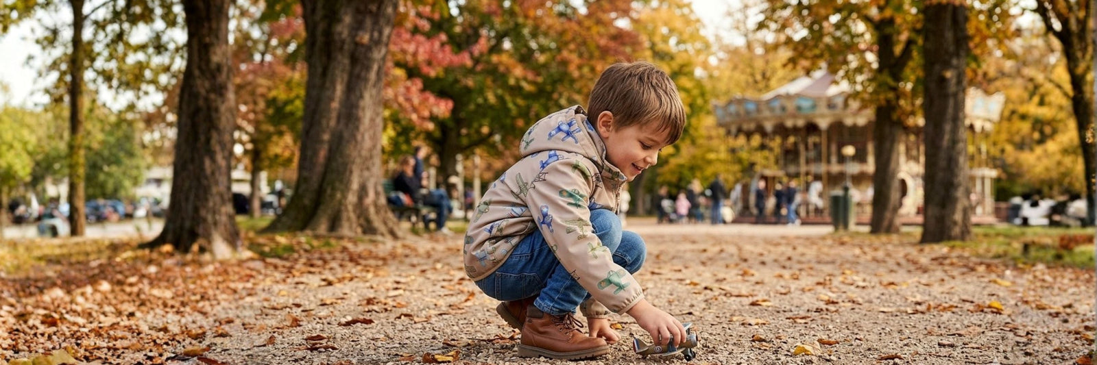 A young boy crouching down to play with a small toy on a gravel path in a park with autumn foliage and a carousel in the background.