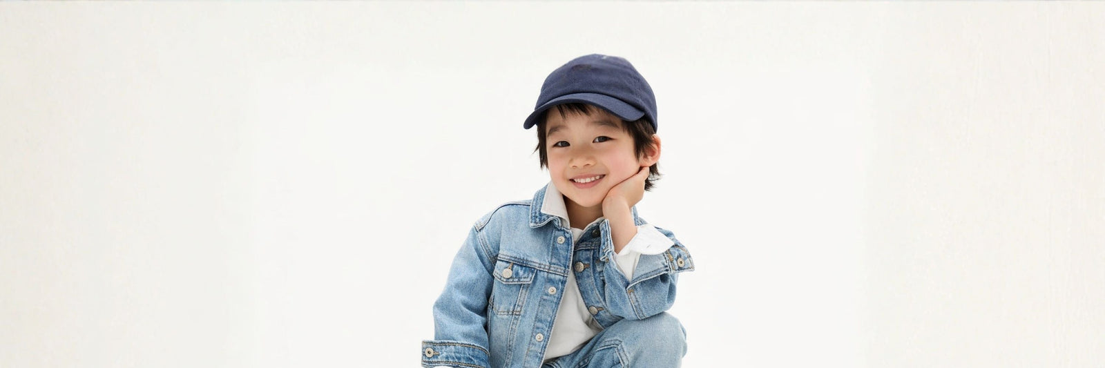A smiling boy in a denim jacket and cap poses playfully with his hand on his cheek against a clean, minimal background.