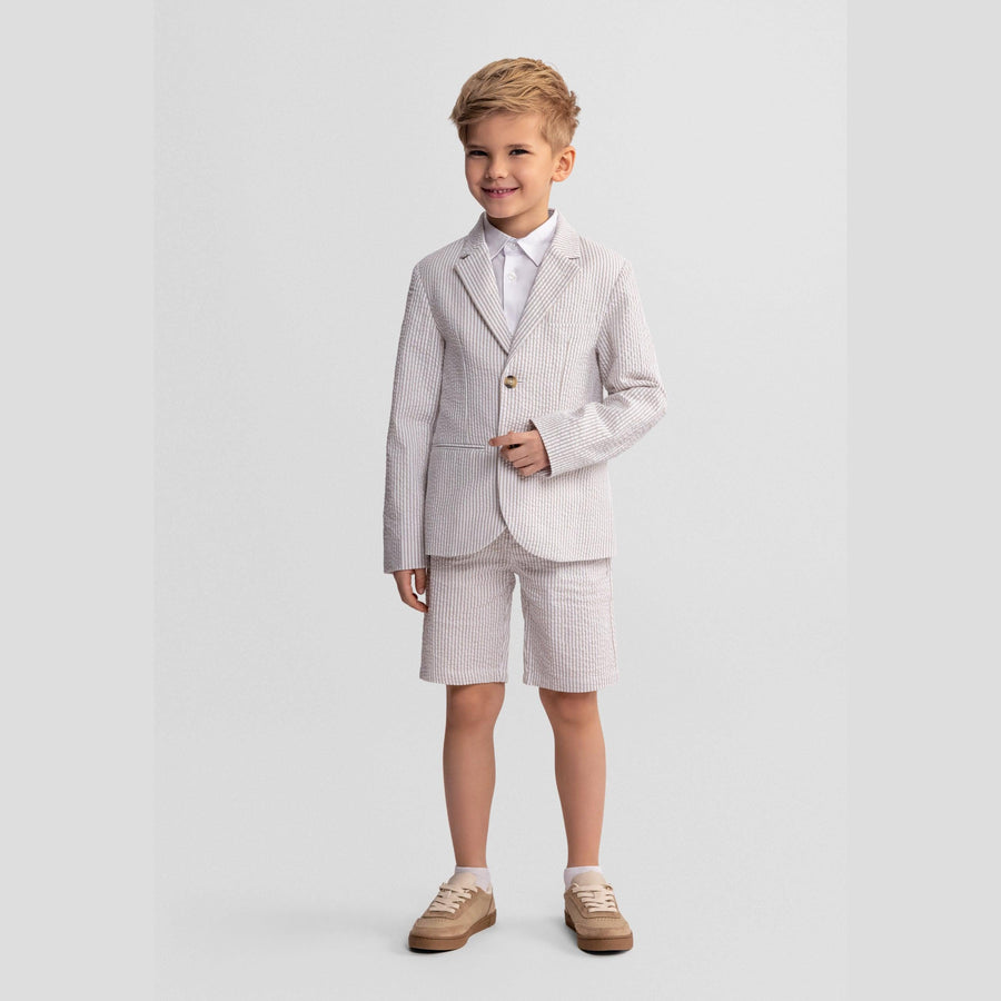 Young boy wearing stone striped cotton blazer with white shirt and matching shorts, standing against white background with hands on hips