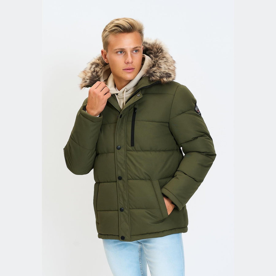 Young male model wearing khaki padded jacket with faux fur hood over cream hoodie and light blue jeans, standing against white background