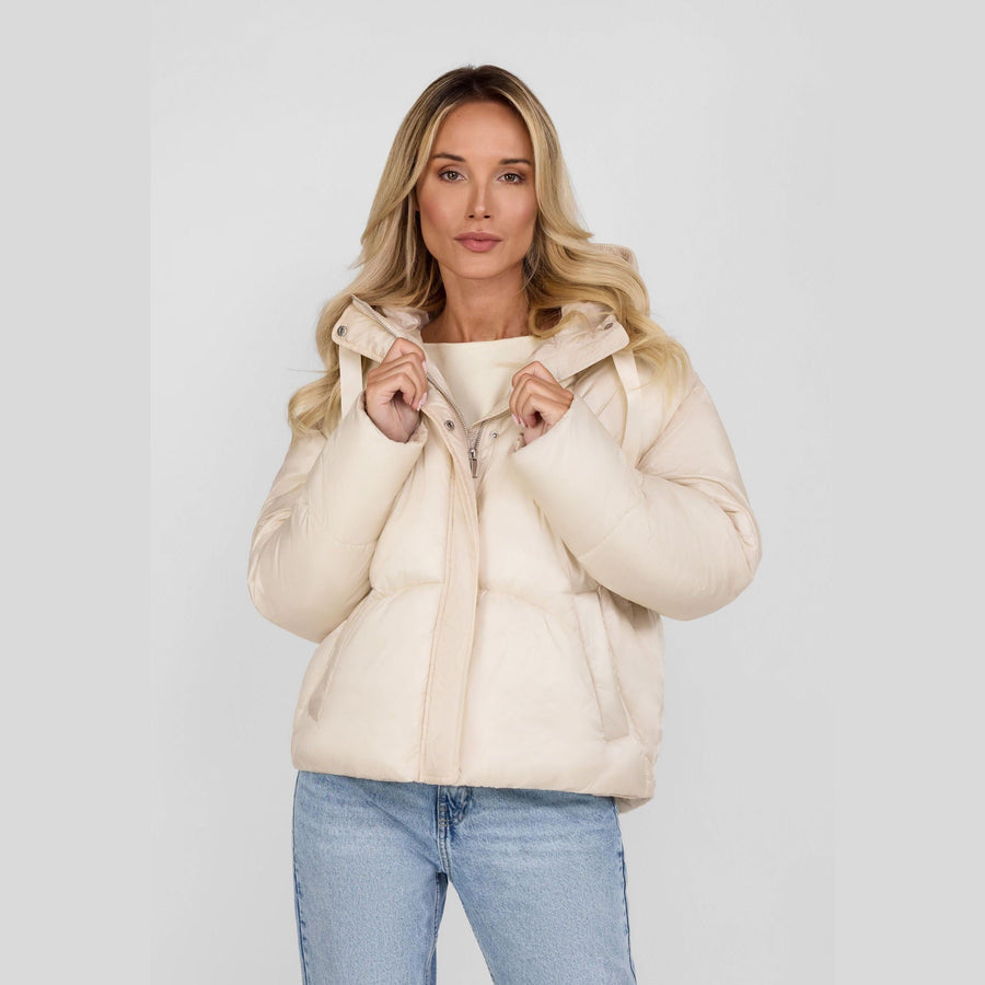 Woman wearing cream puffer jacket with hood up, paired with light blue jeans against white studio background