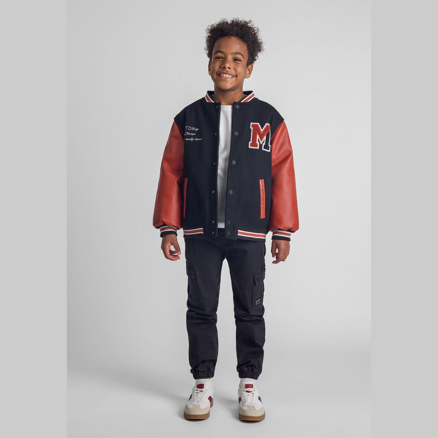 Young boy wearing black and orange varsity jacket with matching dark joggers and white sneakers, standing against light grey background, smiling