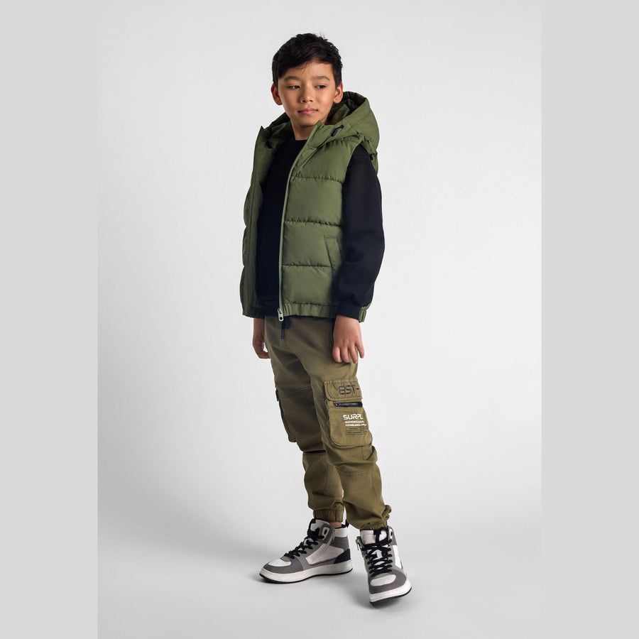 Boy wearing khaki padded gilet with hood over black sweater and matching khaki cargo pants with patches, standing against white background