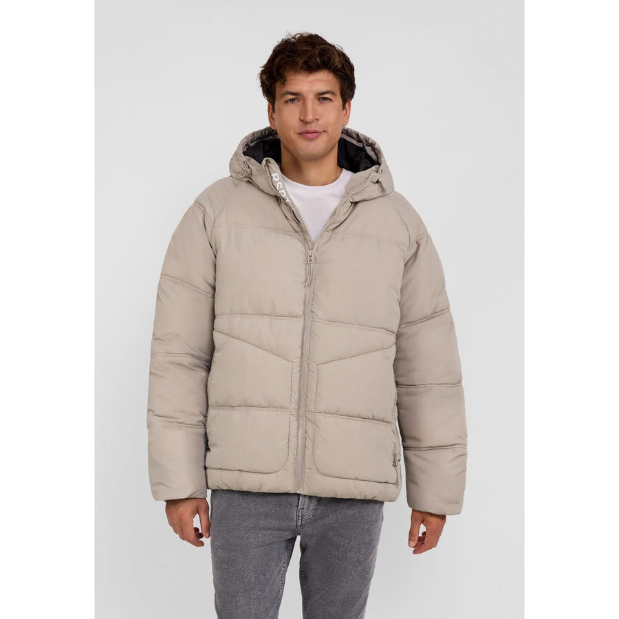 Young male model wearing grey quilted hooded jacket with white t-shirt and grey jeans against light background, front view showing full outfit