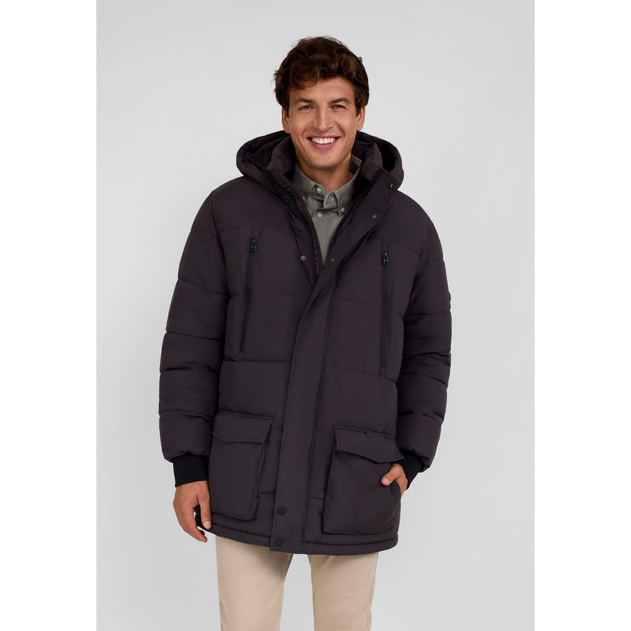 Young man wearing grey padded jacket with hood up, standing against white background, smiling at camera
