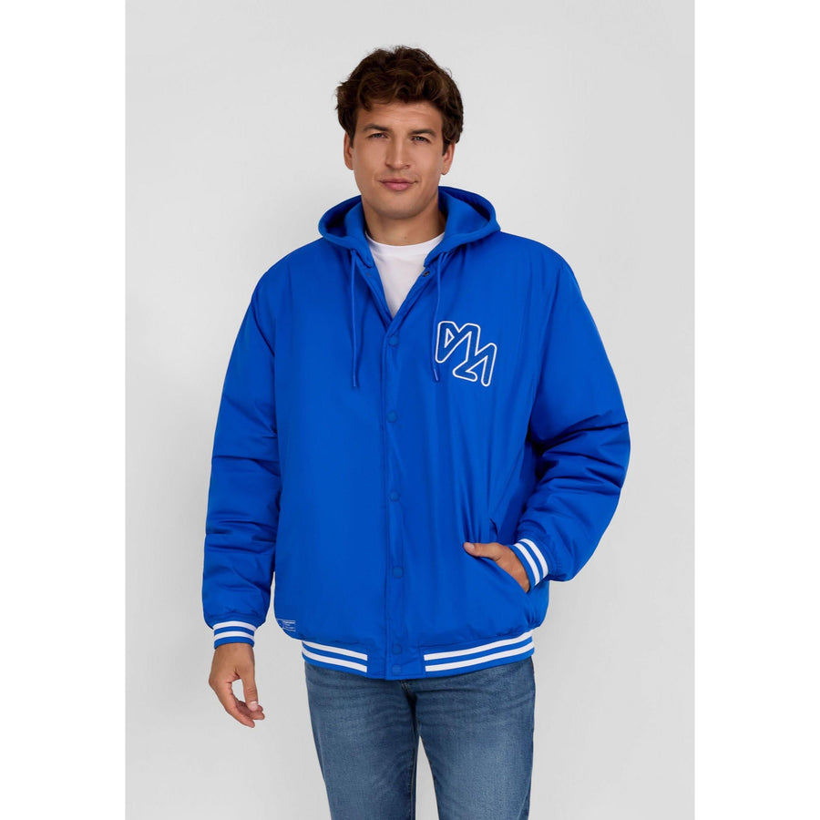 Young man wearing cobalt blue bomber jacket with detachable hood and white t-shirt, hands in pockets, against grey studio background