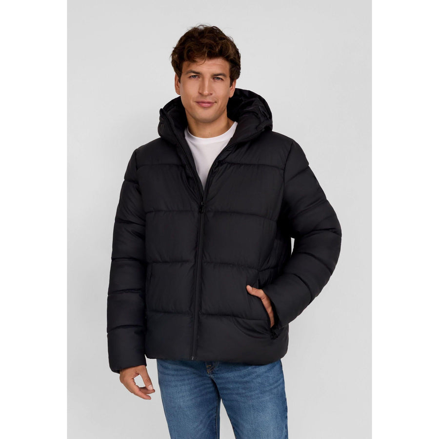 Young man wearing black quilted PU jacket with hood up, hands in pockets, standing against light grey background