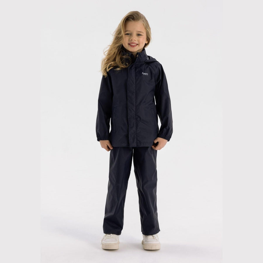 Young girl wearing navy waterproof overpants and matching jacket, standing against white background with blonde hair and white sneakers