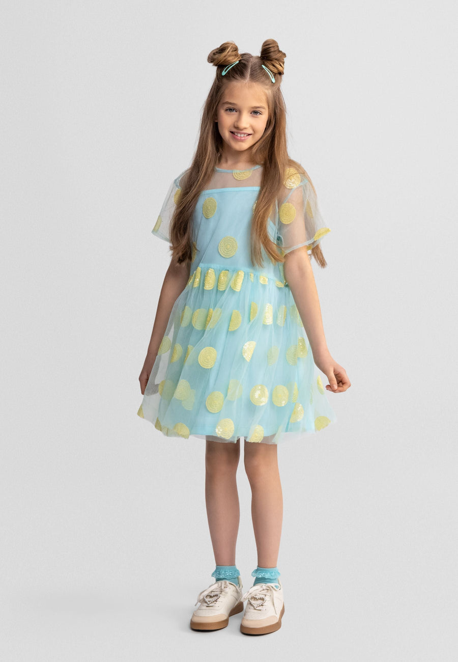 Young girl wearing turquoise tulle party dress with gold sequin polka dots, short sleeves, layered skirt, standing against white studio background