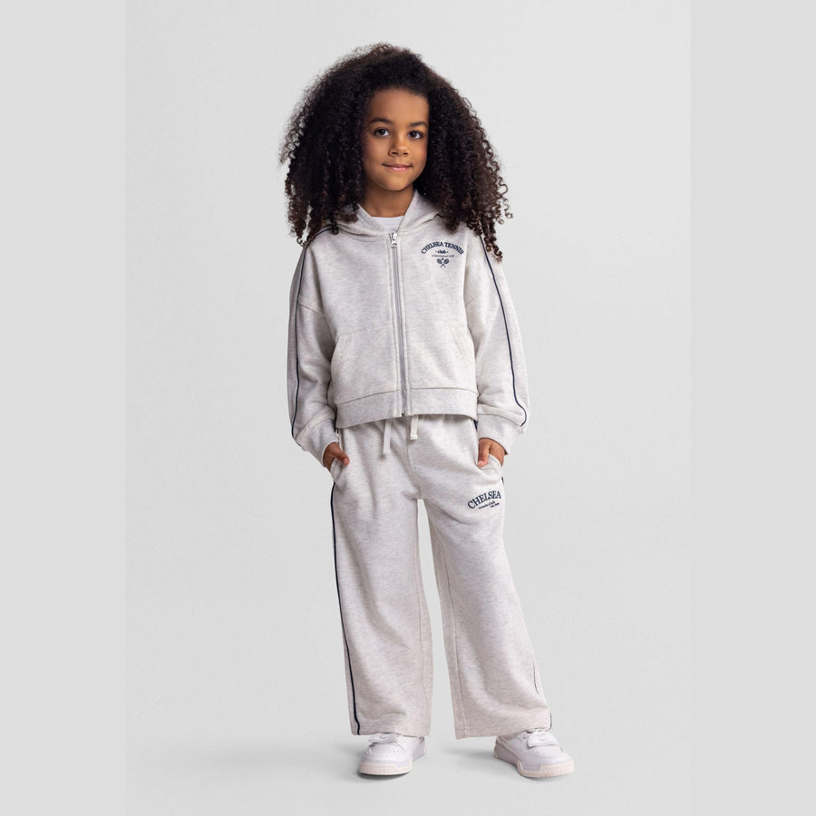Young girl wearing grey marl hooded tracksuit with Chelsea team club branding, standing against neutral background