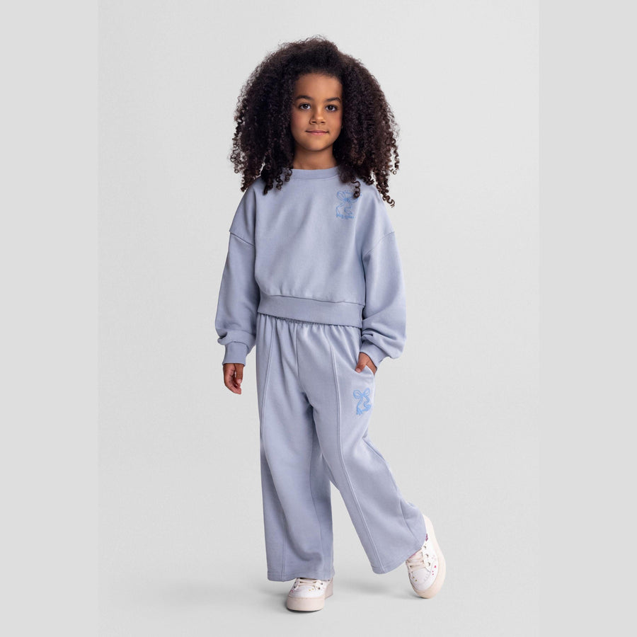 Young girl wearing light blue tracksuit set with wide-leg trousers and cropped sweatshirt, standing against neutral background