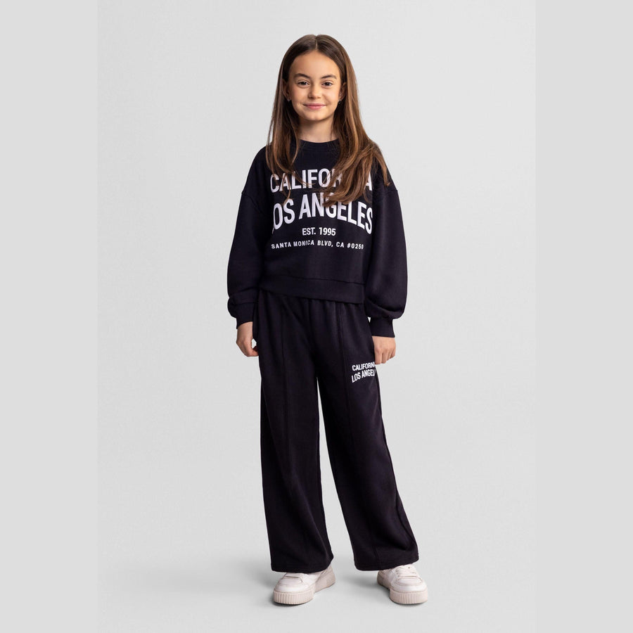 Girl wearing black California Los Angeles tracksuit set with crew neck sweatshirt and loose joggers, standing against light grey background wearing white sneakers