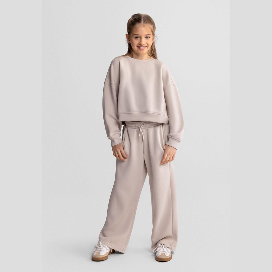 Girl wearing beige wide leg joggers with matching sweatshirt, standing against white background, showing full outfit coordination