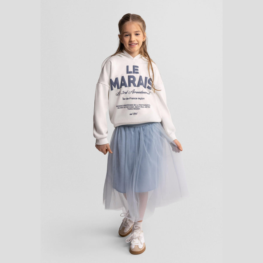 Young girl wearing grey pleated tulle maxi skirt with white hoodie, standing against white background, full body shot showing the flowing movement of the skirt