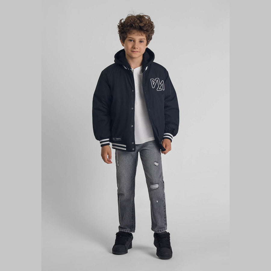 Boy wearing black baseball jacket with detachable hood over white t-shirt and distressed grey jeans, standing against light grey background