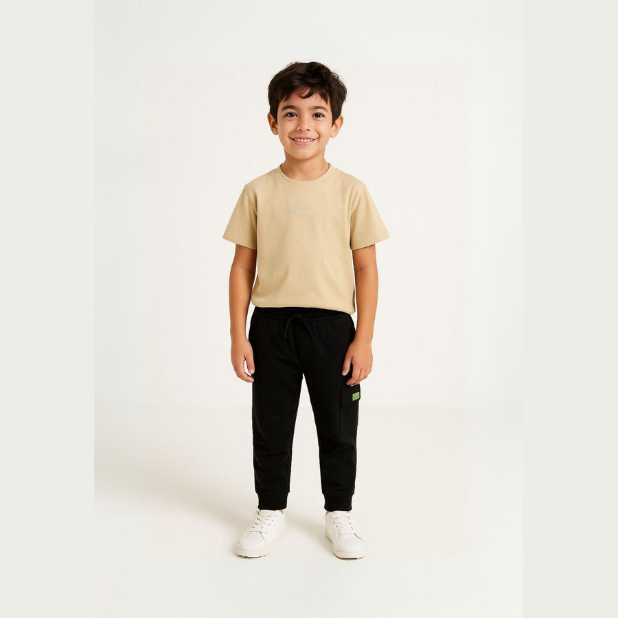 Young boy wearing sand coloured t-shirt with black joggers standing against white background