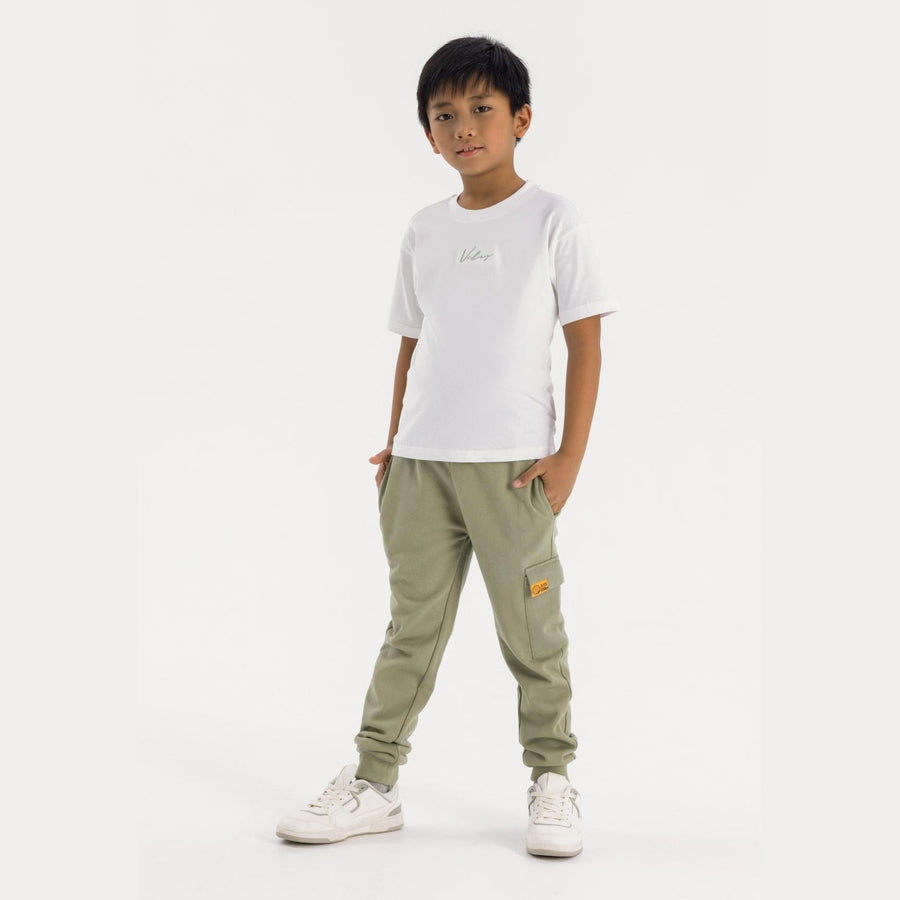 Young boy wearing olive green t-shirt and joggers set, standing against white background with hands in pockets