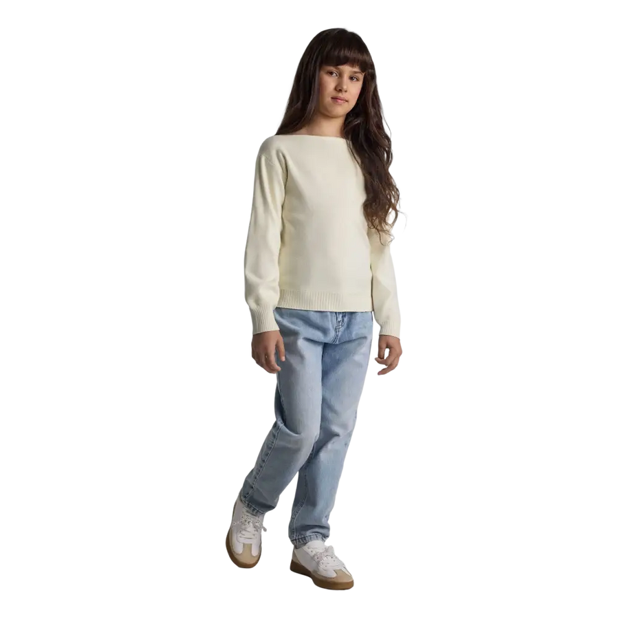 Young girl wearing cream textured knit jumper with boat neckline and light blue jeans, standing against black background with long brown hair
