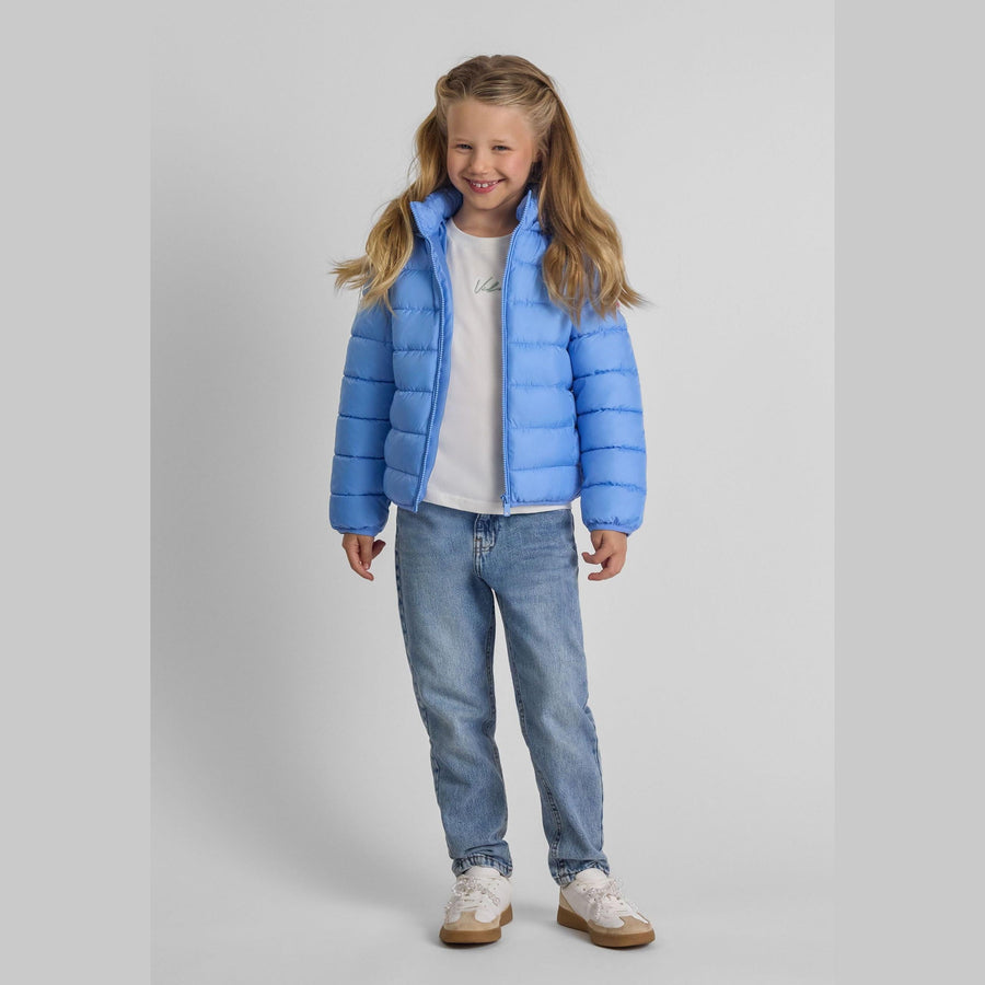Young girl wearing blue quilted jacket with detachable hood, white top and jeans, smiling against light grey background