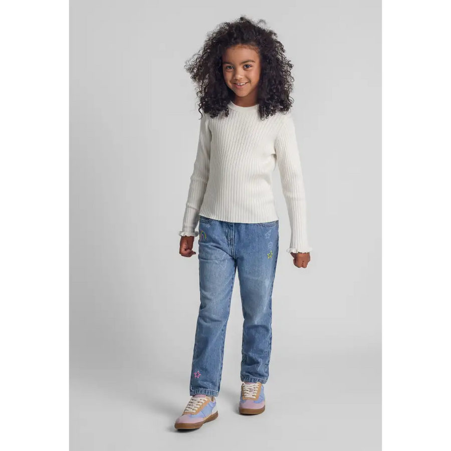Young model wearing white ribbed jumper with frill cuffs paired with blue jeans, full body shot on light grey background