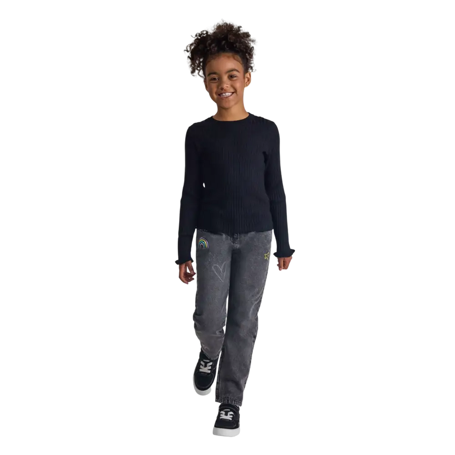 Young girl wearing black ribbed jumper with frill cuffs and grey jeans, full body shot on black background